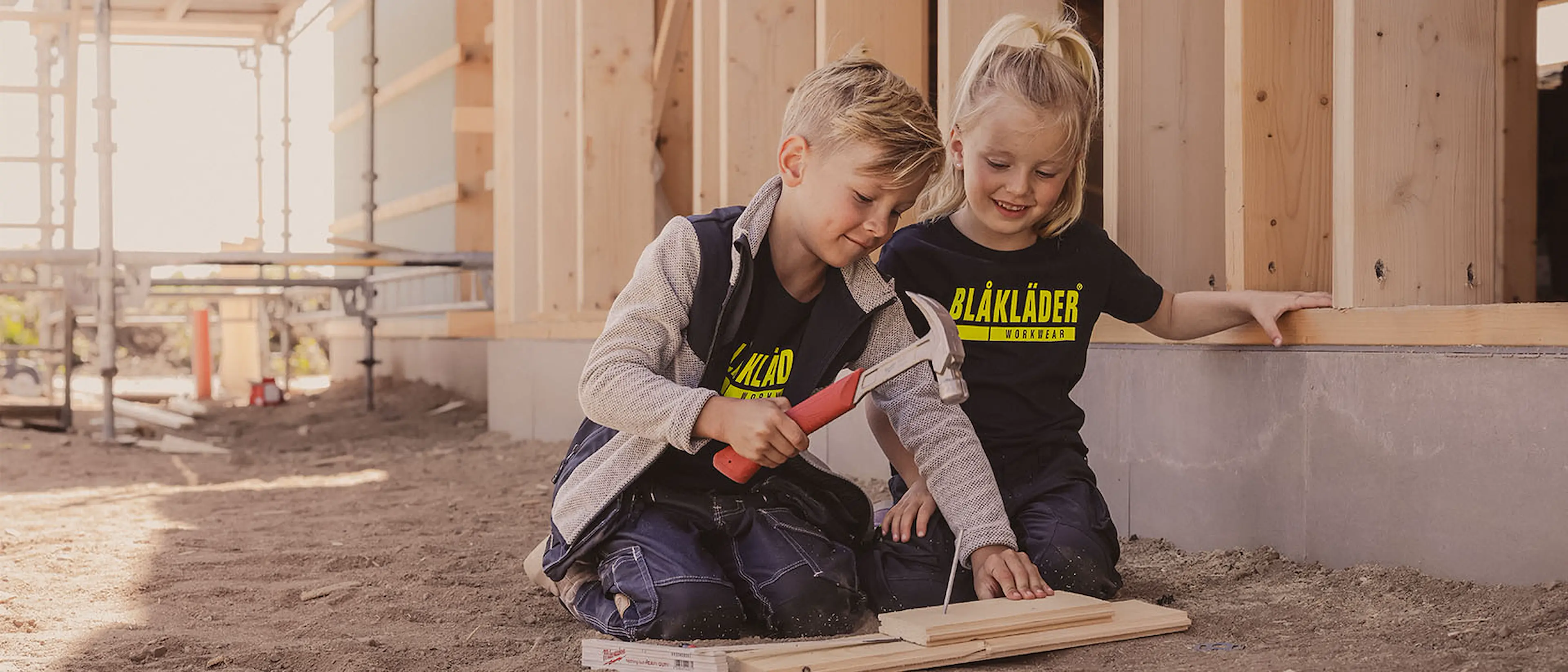 Två barn i Blåkläder arbetskläder sitter på en byggarbetsplats och spikar trä tillsammans med en hammare.