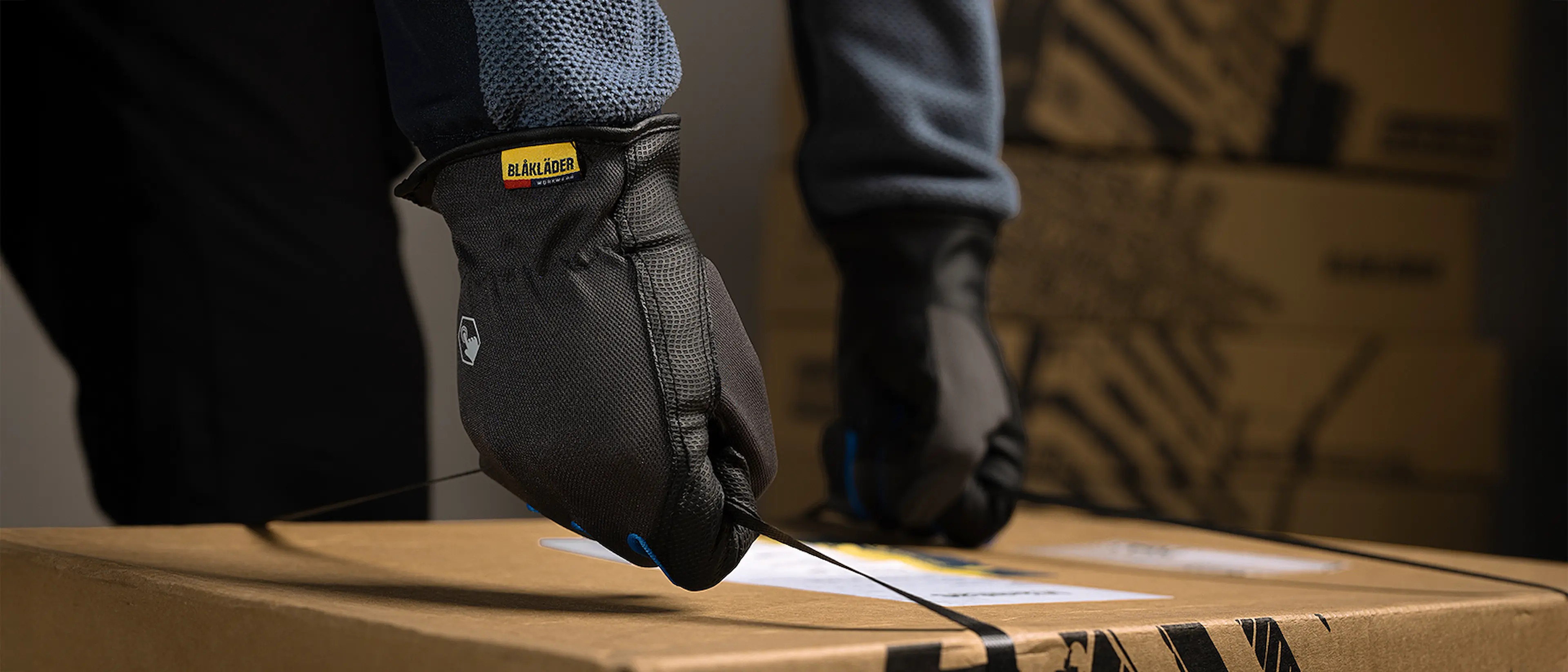 Close-up of gloved hands tightening plastic straps around a cardboard box, with stacked boxes in the background.