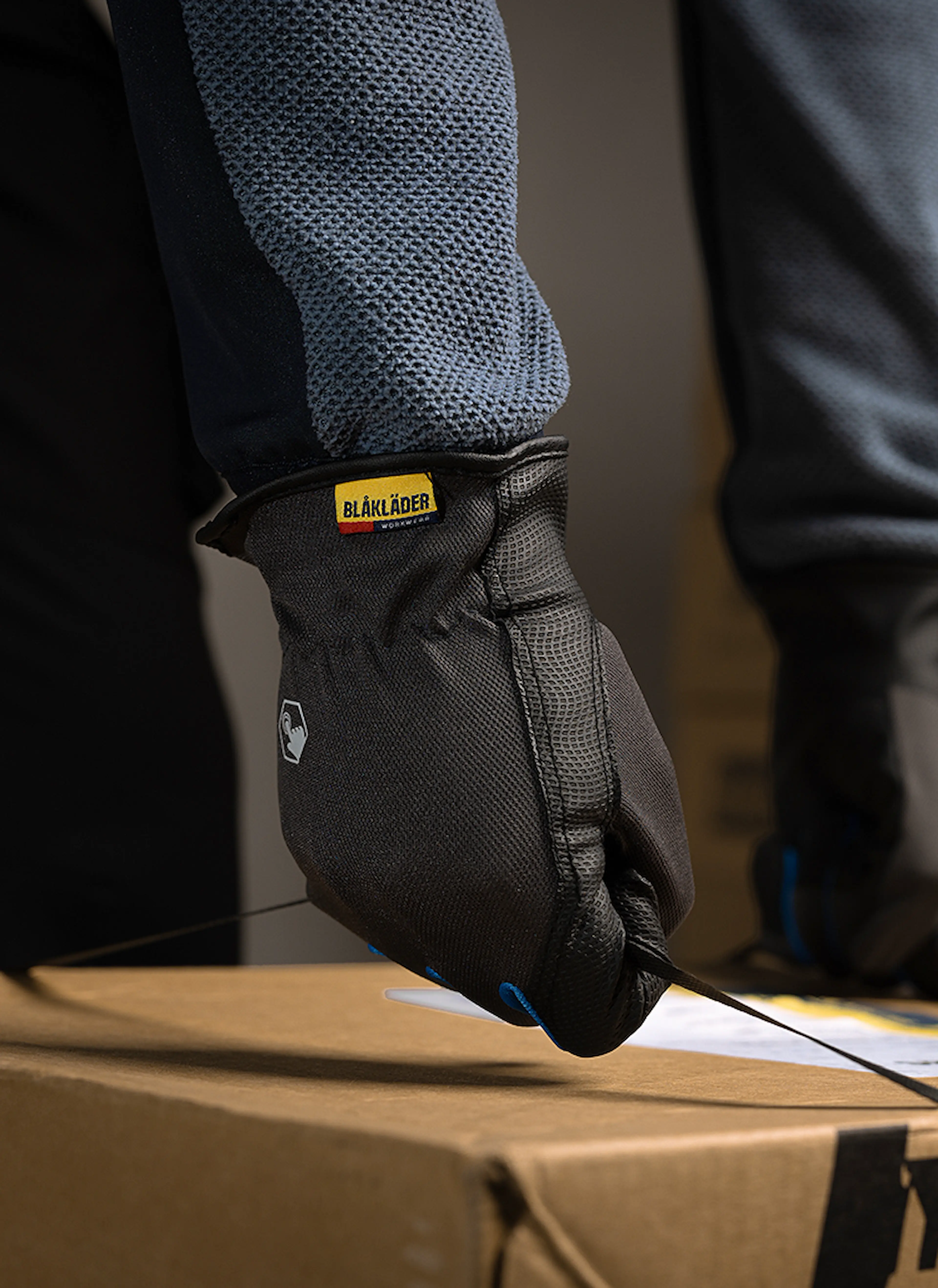 Close-up of gloved hands tightening plastic straps around a cardboard box, with stacked boxes in the background.